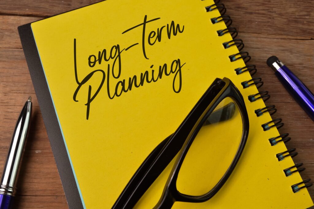 Glasses on top of a yellow notebook that says long-term planning on top of a table next to two pens