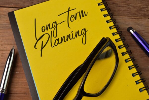 Glasses on top of a yellow notebook that says long-term planning on top of a table next to two pens